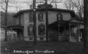 Historic two-story house with ornate architecture, surrounded by trees.