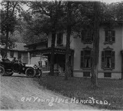 Historic homestead with a vintage car parked in front, surrounded by trees.