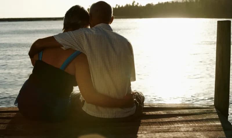 A couple sits together on a dock, embracing as the sun sets over the water.