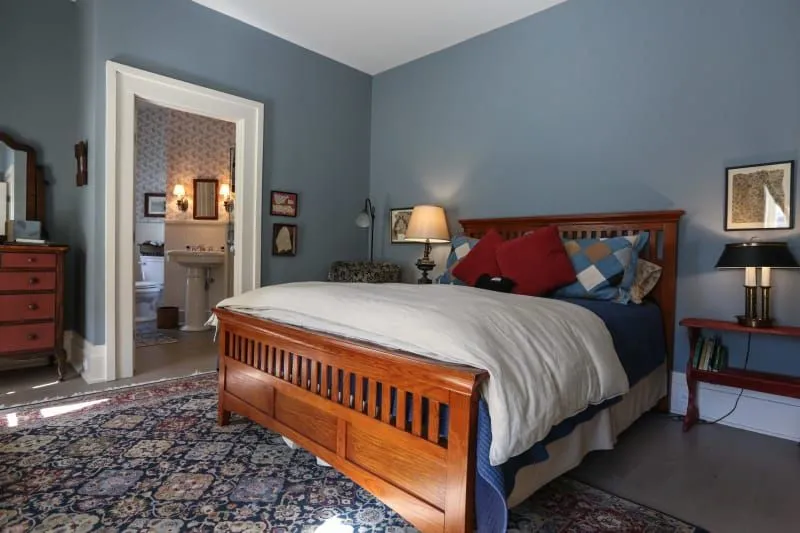 A cozy bedroom featuring a wooden bed, colorful pillows, a side table with a lamp, and a doorway leading to a bathroom.