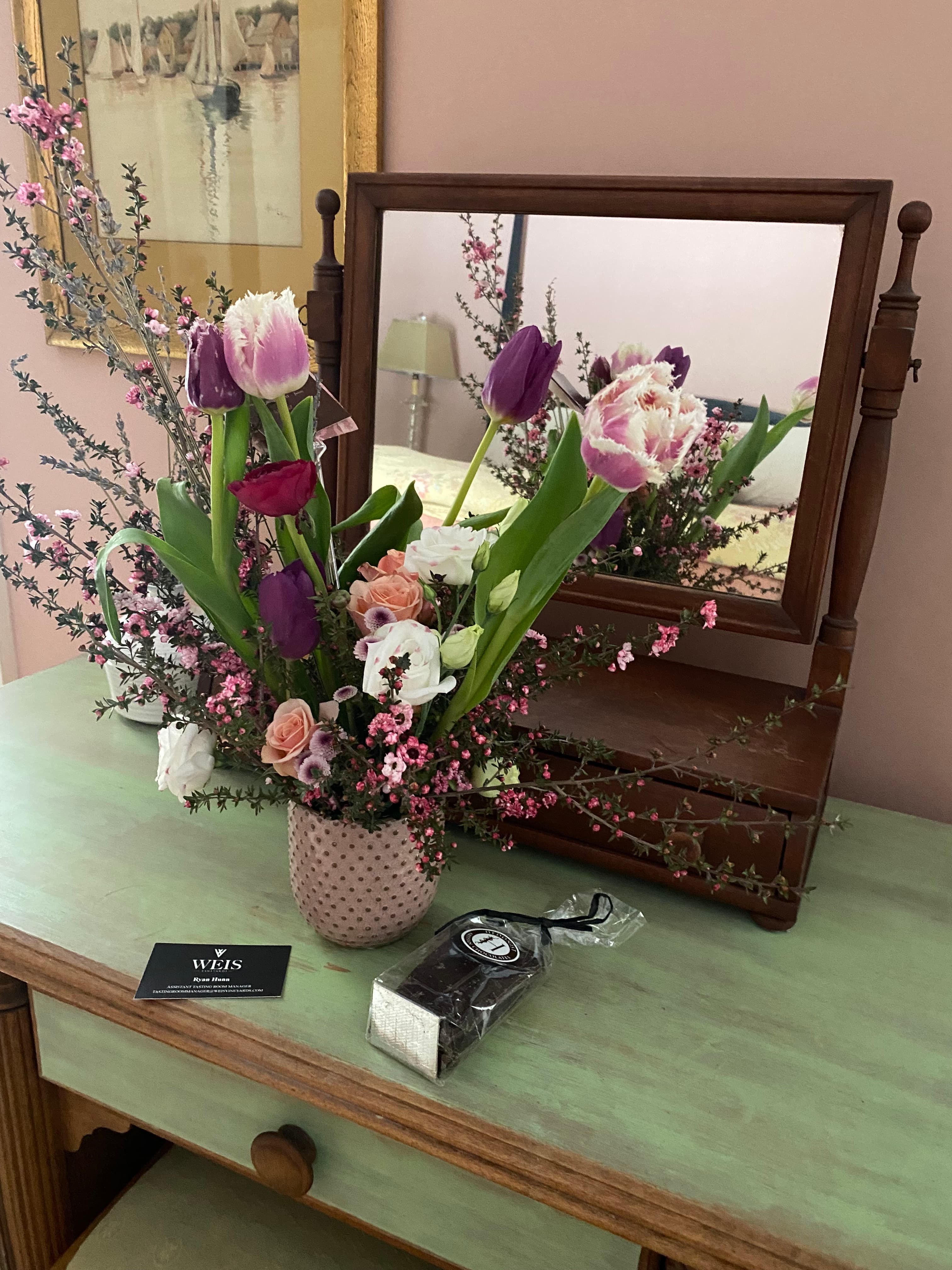 A vibrant floral arrangement in a pink pot is displayed on a green dresser beside a small box of chocolates and a mirror.