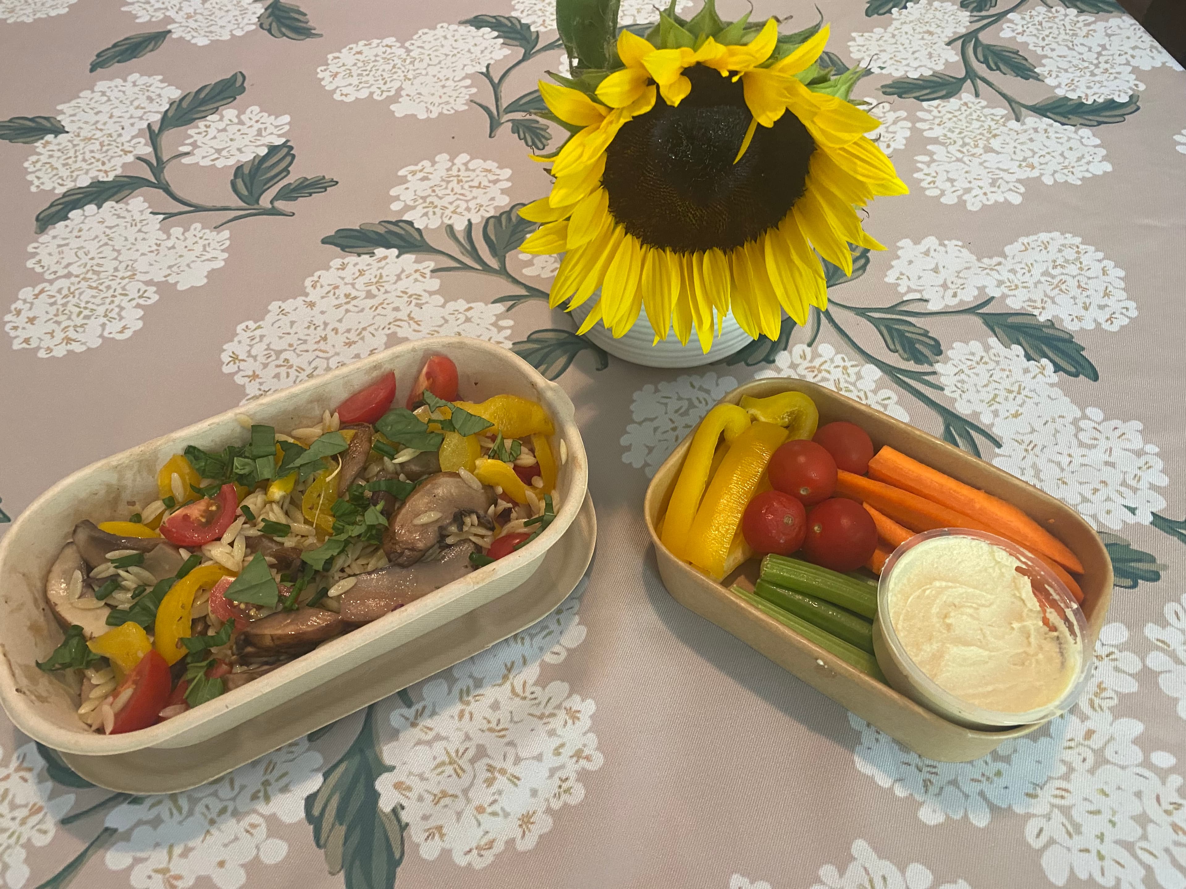 A bowl of colorful vegetable stir-fry and a platter of fresh veggies with hummus, beside a vibrant sunflower on a floral tablecloth.