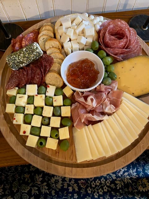 A wooden platter featuring an assortment of cheeses, cured meats, olives, crackers, and a small bowl of jam.