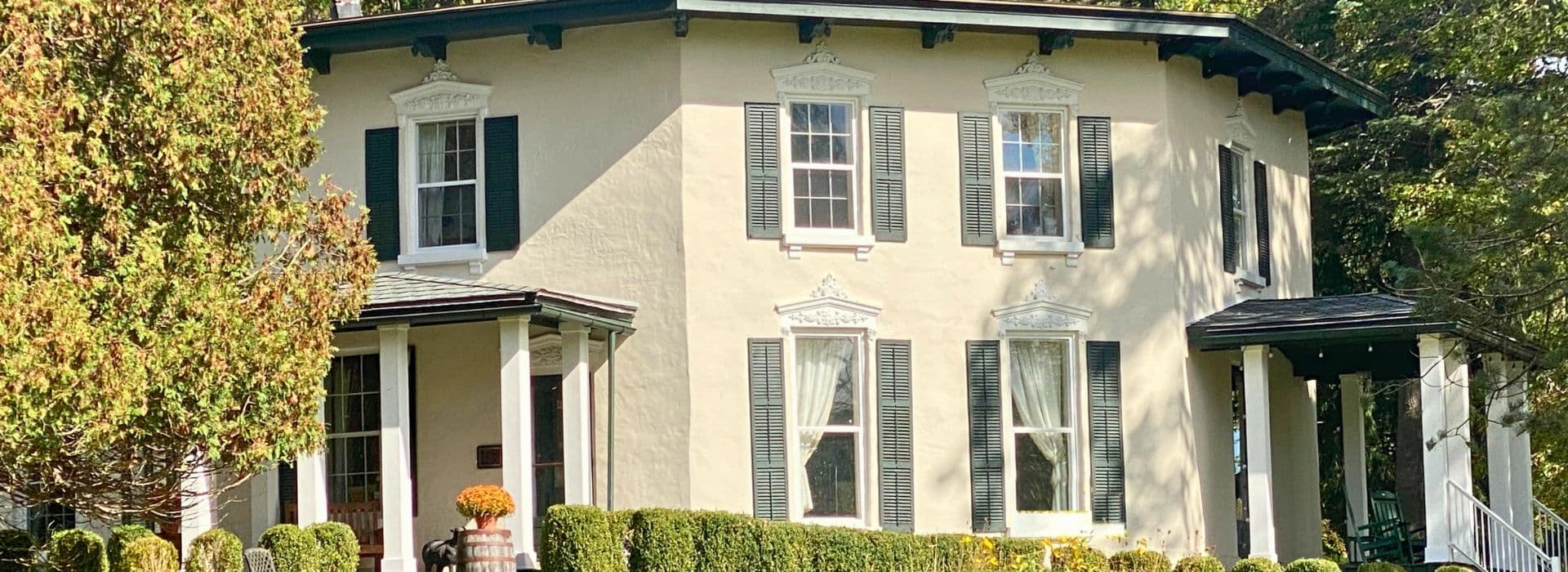 A charming two-story house with green shutters and a circular design, surrounded by neatly trimmed bushes.
